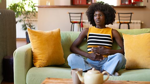 A woman sits on the sofa holding her stomach in pain 