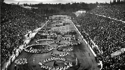 People are grouped in the shape of the Olympic rings in a crowded stadium. 