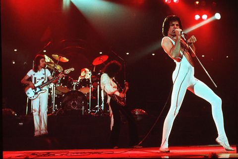 Two men playing electric guitars on stage stood in front of a drum kit.  At the front of the stage is a singer dressed in a white unitard singing into a portable microphone.