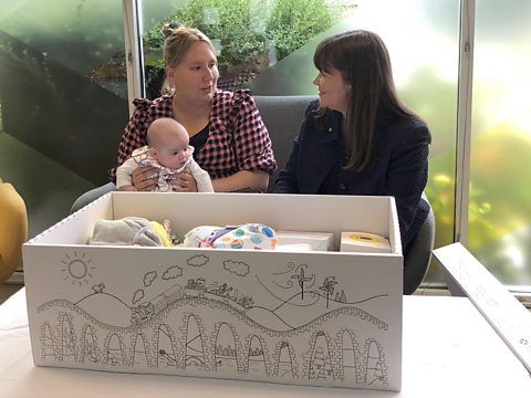 A mother with a babying a woman. In front of them is a cardboard baby box containing baby items.