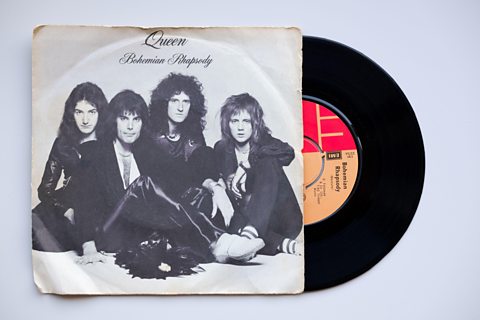 The record sleeve for Queen's Bohemian Rhapsody with 4 band members posed sat casually on the floor.  A record can be seen half removed from the sleeve. 