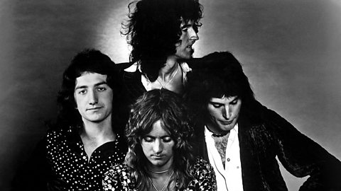 A black and white image of four male performers posed in a group, one is looking at the camera the others are looking down and to the right.  All with long hair wearing partially buttoned or open shirts. 