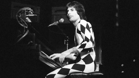 A black and white image of a male performer on stage wearing a tight all in one suit with large black and white diamonds on it.  He is playing a piano whilst singing into a microphone.