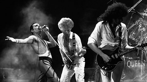 Black and white photo of three men performing on stage.  Two are playing electric guitars and the third is holding a portable mic to his mouth and walking whilst singing with his right arm extended towards the audience..