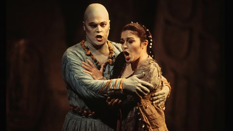 Two operatic performers on a stage with a black background, embracing whilst singing.