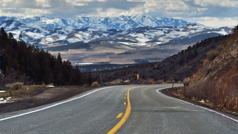 A winding road with yellow road markings down the centre.  To either side their are trees and shrubbery and snow capped mountains against a cloudy sky in the horizon.