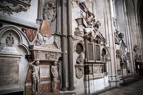 Poets’ Corner in Westminster Abbey, wall of stone memorial plaques surrounded by a mixture of statues and coats of arms