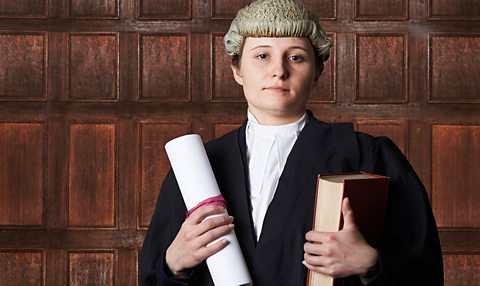 A female barrister wearing a wig holds a book and a scroll in a courtroom