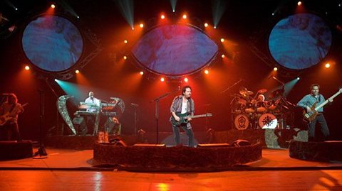 A picture of Toto in concert.  Four male musicians two playing guitar, one on keyboard and one on drums.  They are all stood on separate raised areas of the stage and the lighting is red.