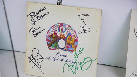 An album record cover with a colourful circular pattern in the centre and the text Queen A Night at the Opera below. In the surrounding white space there are 4 signatures of the band members and a handwritten dedication pf To Darlene, from Queen.