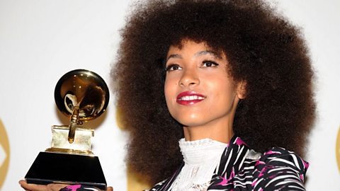 A woman wearing a white high collared blouse and a zebra print jacket with pink accents.  She is smiling and holding a trophy of a gramophone in her right hand.