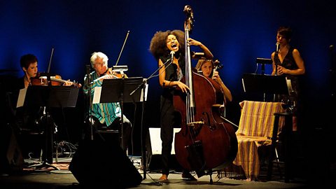 A group of musicians on stage playing a violin, viola, cello and percussion behind a bassist who is singing at the front of the stage.