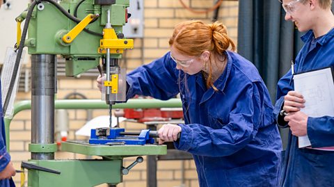 Apprentice and colleague in blue overalls using a drill.