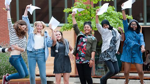 Students in front of a school holding their results papers up in the air in celebration.