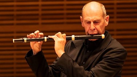 A man dressed in a black shirt and suit sat on stage playing a Baroque flute.