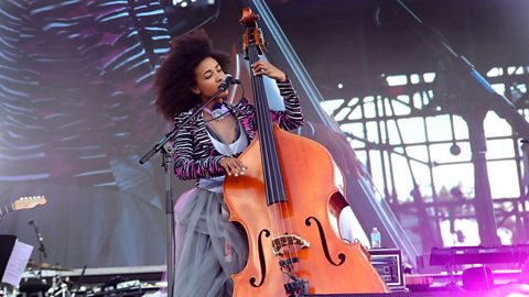 A woman wearing a zebra print jacket and white top playing a double bass, singing into a microphone performing on an outdoor stage. 