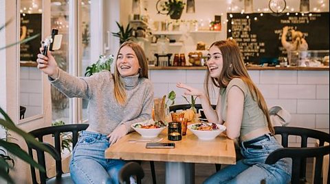 Image of 2 girls in a coffee shop taking a selfie with their food