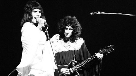 A black and white photo of two male performers.  One is playing an electric guitar and the other one is singing into a stand up microphone.