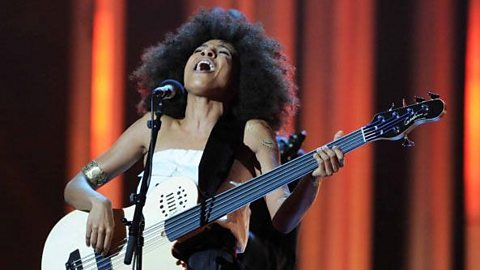 A female singer on stage playing a bass guitar.