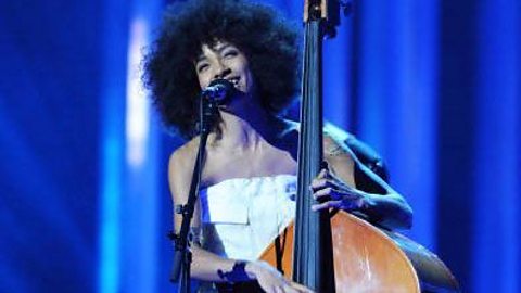 A female musician performing on a stage playing an upright bass and singing into a freestanding microphone.