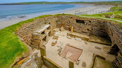 An aerial view of the inside of a ruin of a stone house, on a beach, with stone furniture visible