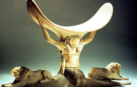 An ornate curved headrest supported by a column and plinth shaped in the figures of lions and an Egyptian individual 