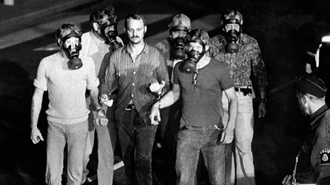 Getty Images Men in gas masks handcuffed to central man, with police officer looking from the side (Credit: Getty Images)