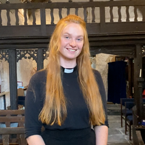 A young woman with long ginger hair, wearing black with a white clerical collar, stands with hands clasped in a church