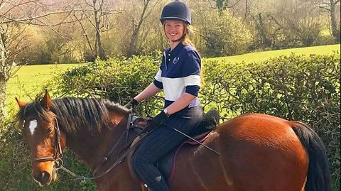 Young woman wearing navy and white clothes and a navy riding helmet rides a brown horse