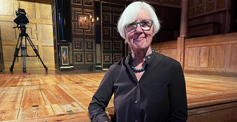 Photograph of composer Judith Weir, facing the camera and smiling, whilst leaning with one elbow on the stage of The Globe Theatre