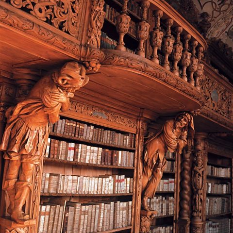 Two wooden carvings of hunched over men stand either side of a bookshelf, supporting the carved wooden balcony above on their shoulders