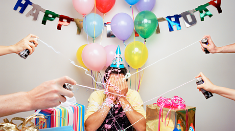 Someone sits with their hands covering their eyes. They are being spray with silly string. They are surrounded by presents, balloons and a sign that reads 'happy birthday'.