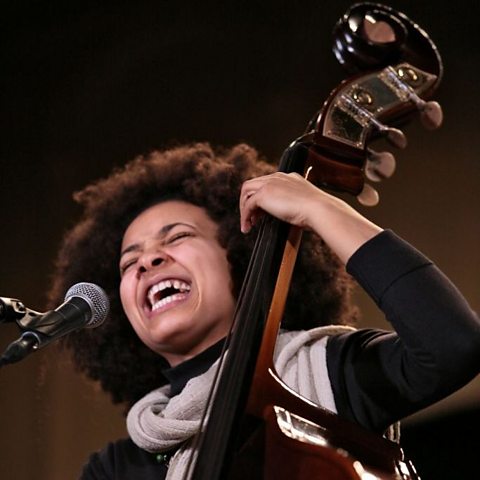 A photo of a female singing into a microphone and playing a double bass.