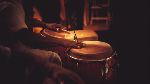 Hands of a person who is sat playing tall conga drums between their legs.