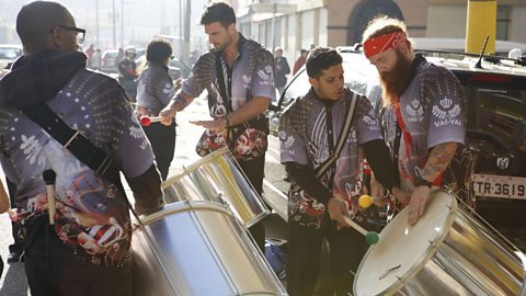 A group of four men in a street parade wearing large silver drums around their torsos and beating them with drumsticks with coloured ball ends.