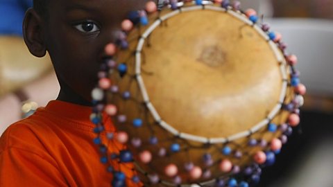 A young boy holding a percussion instrument made from dried gourd and surrounded by beads woven into a net.