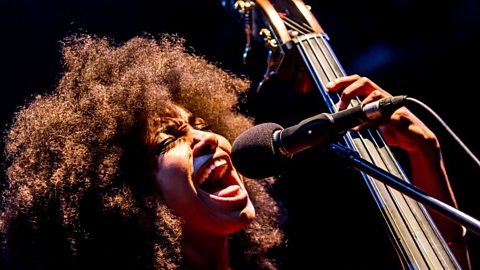 A close up photo of the head of a female singing into a microphone whilst playing a double bass. 