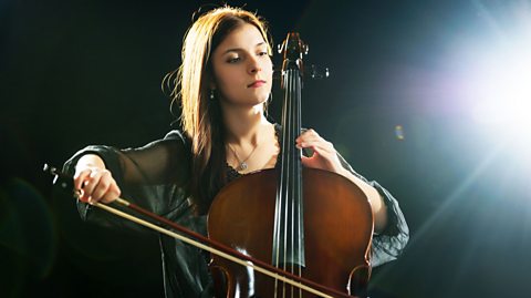 A photo of a female musician playing a cello with a black background and a spotlight from the right.