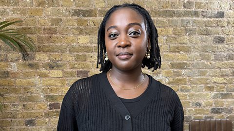 Photo of composer Cassie Kinoshi, posing with a smile in front of a trendy brickwall