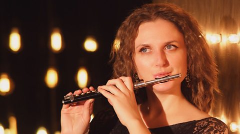 A head shot picture of a female with red hair playing a piccolo.  There are rows of warm yellow lights in the background.