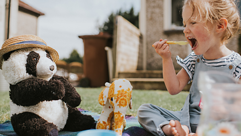 A young girl hosts a teddy bear's picnic, lifting a spoon of food to her mouth, with a plush elephant and a toy panda wearing a straw boater hat.