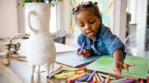 A young girl picks out her next crayon colour, while drawing on coloured paper. There is a precarious vase in the foreground.