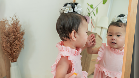 A young girl wearing a pink top looks at her reflection in a mirror.