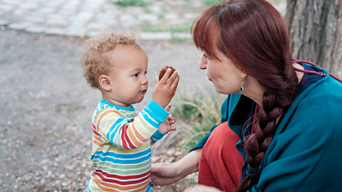 A toddler in a colourful stripey jumper shows an object he has found in the park to his mum, who crouches down to his level.