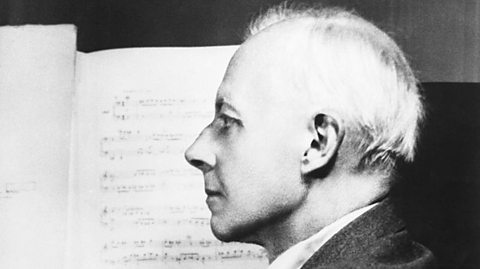 A black and white photo of a white older man in profile, sat at a piano with sheet music behind him.