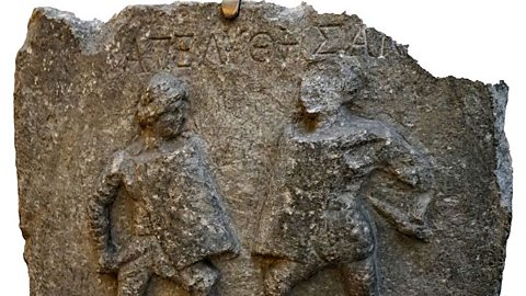 A square of marble with two sculpted women, facing each other to fight, and Ancient Greek writing above them.