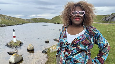 Composer Errollyn Wallen smiling, standing next to a small lake with rocks and a mini lighthouse on one of the rocks