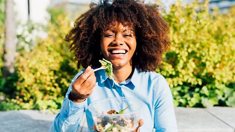 A woman eats a chicken salad while standing outside smiling 