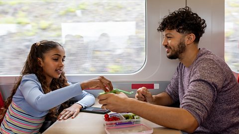 A side-view shot of a smiling father and his young daughter sitting opposite each other on a train journey. They are both leaning on the table, the young girl taking a piece of popcorn from the plastic container which her father is offering to her. 