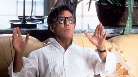 Dustin Hoffman sits on a beige coach with his arms raised in gesture, wearing large glasses and a white shirt